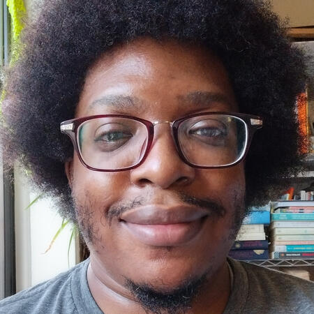 A Black man with an afro is wearing a gray shirt and smiling while standing next to a window. Piles o books are visible on the shelf behind him.